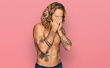 Handsome man with beard and long hair standing shirtless showing tattoos rubbing eyes for fatigue...