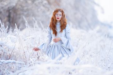 Young redhead woman, a princess, walks in a winter forest in a blue dress.