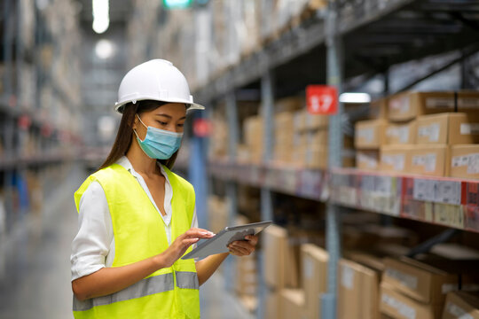 The Working During Outbreak Of Covid-19 Concept: Female Warehouse Staff With Face Mask Use Tablet Check Goods In Warehouse.