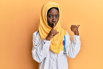 Beautiful african young woman wearing doctor uniform and hijab asking to be quiet with finger on lips pointing with hand to the side. silence and secret concept.