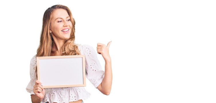 Young caucasian woman with blond hair holding blank empty banner pointing thumb up to the side smiling happy with open mouth