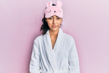 Hispanic teenager girl with dental braces wearing sleep mask and robe with serious expression on face. simple and natural looking at the camera.