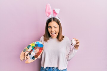 Young beautiful woman wearing cute easter bunny ears holding painting palette and egg celebrating crazy and amazed for success with open eyes screaming excited.