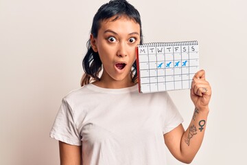Young woman holding travel calendar scared and amazed with open mouth for surprise, disbelief face