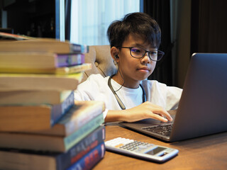 Closeup face of an Asian boy wearing glasses and earphone doing home study with stack of books in the foreground.
