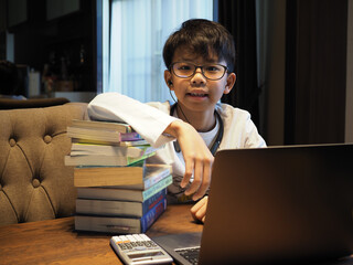 Closeup face of an Asian boy wearing glasses and earphone looking at the camera. He is studying at home with stack of books. Foreground have a silver laptop.