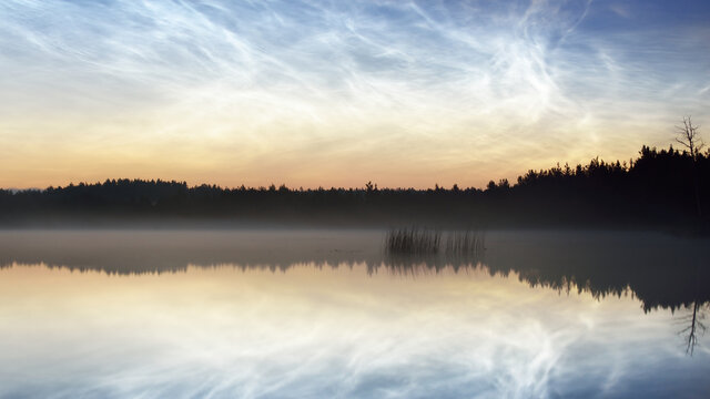 Starry sky with noctilucent clouds and fog above the Saimaa lake, Finland, at summer solstice night. Long exposure. Symmetry reflections, natural mirror. Mystic landscape, pagan celebration