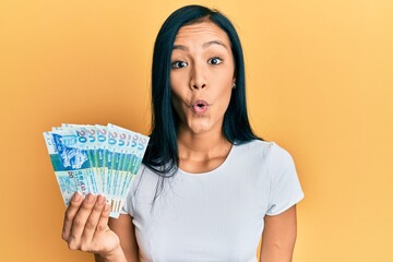 Beautiful hispanic woman holding hong kong 20 dollars banknotes scared and amazed with open mouth for surprise, disbelief face