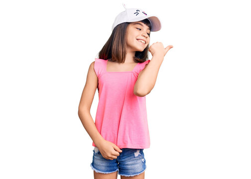 Young little girl with bang wearing funny kitty cap smiling with happy face looking and pointing to the side with thumb up.