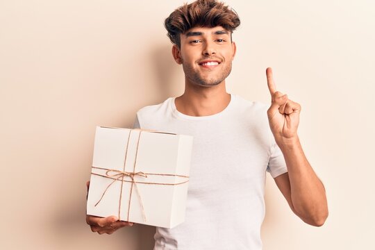 Young hispanic man holding gift smiling with an idea or question pointing finger with happy face, number one