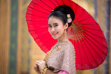 Beautiful woman in a pink Thai dress or a traditional Thai dress is spreading a red umbrella in a Thai temple.