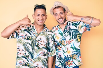 Young gay couple of two men wearing summer hat and hawaiian shirt smiling doing phone gesture with hand and fingers like talking on the telephone. communicating concepts.