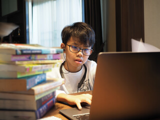 Closeup face of an Asian boy wearing glasses and earphone doing home study with stack of books in the foreground.