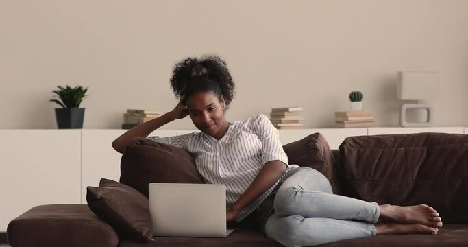 Young African woman relaxing on comfy sofa in living room alone use portable computer watching videos in social media networks, tv series on internet, spend lazy weekend time using modern tech concept