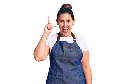 Young beautiful brunette woman wearing apron pointing finger up with successful idea. exited and happy. number one.