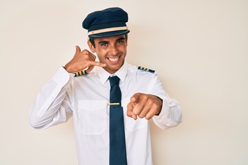 Young hispanic man wearing airplane pilot uniform smiling doing talking on the telephone gesture and pointing to you. call me.