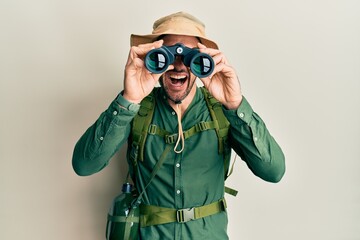 Handsome man with beard wearing explorer hat looking through binoculars smiling and laughing hard out loud because funny crazy joke.