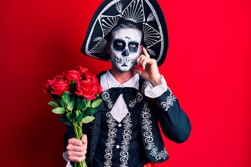 Fototapeta premium Young man wearing mexican day of the dead costume holding roses serious face thinking about question with hand on chin, thoughtful about confusing idea