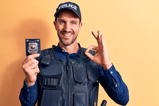 Young handsome policeman wearing uniform and bulletprof holding police badge doing ok sign with fingers, smiling friendly gesturing excellent symbol