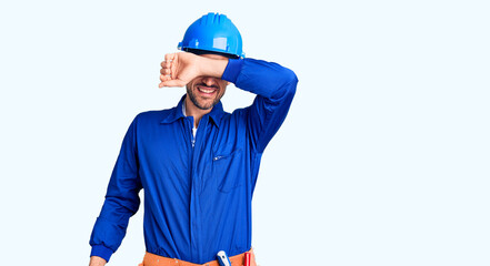 Young handsome man wearing worker uniform and hardhat covering eyes with arm smiling cheerful and funny. blind concept.