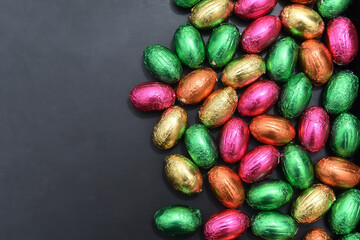 Foil wrapped multi coloured easter eggs in pink, green, orange and yelow in a pile or group, against a grey black background.