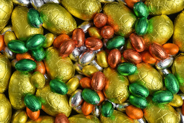 Large & small yellow, gold, orange, green and silver spring colours of foil wrapped chocolate easter eggs, against a black background.