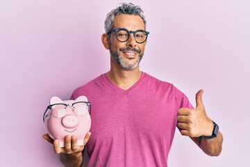 Middle age grey-haired man holding piggy bank with glasses smiling happy and positive, thumb up...