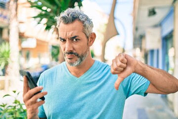 Middle age grey-haired man using smartphone at street of city with angry face, negative sign...