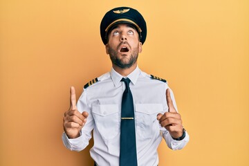 Handsome hispanic man wearing airplane pilot uniform amazed and surprised looking up and pointing...