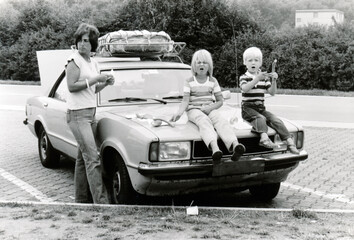 Vintage monochrome 1978 image; young mother with daugter and son sitting on hood of car eathing...