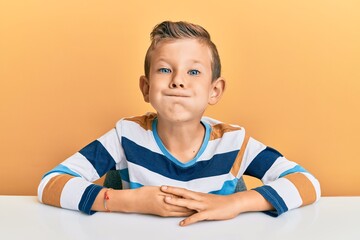 Adorable caucasian kid wearing casual clothes sitting on the table puffing cheeks with funny face....