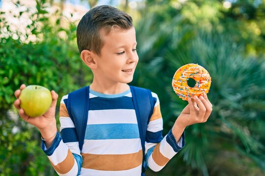 Adorable caucasian student boy smiling happy holding donut and bottle of water at the park.