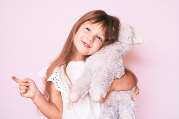 Little caucasian kid girl with long hair holding stuffed animal smiling happy pointing with hand...