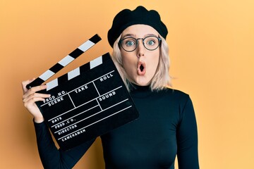 Young blonde girl holding video film clapboard scared and amazed with open mouth for surprise,...