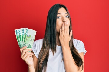 Young brunette woman holding 1000 south korean won banknotes covering mouth with hand, shocked and afraid for mistake. surprised expression