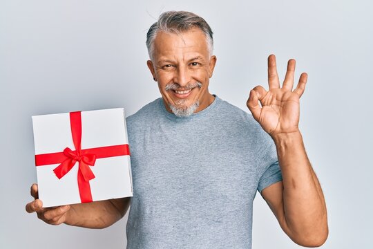 Middle age grey-haired man holding gift doing ok sign with fingers, smiling friendly gesturing excellent symbol