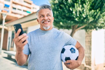 Middle age hispanic grey-haired man using smartphone and holding soccer ball at the city.