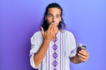 Young handsome man with long hair drinking mate infusion covering mouth with hand, shocked and afraid for mistake. surprised expression