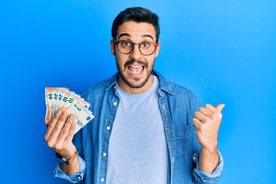 Young hispanic man holding euro banknotes pointing thumb up to the side smiling happy with open mouth
