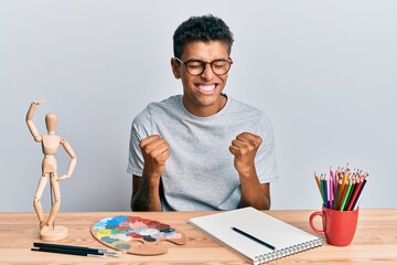 Naklejka premium Young handsome african american man painter sitting palette and art manikin very happy and excited doing winner gesture with arms raised, smiling and screaming for success. celebration concept.
