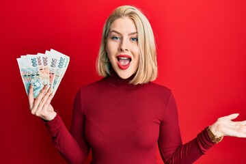 Young blonde woman holding czech koruna banknotes celebrating achievement with happy smile and winner expression with raised hand