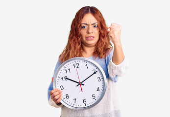 Young latin woman holding big clock annoyed and frustrated shouting with anger, yelling crazy with anger and hand raised