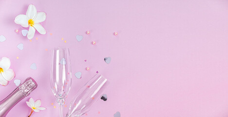 Two empty champagne glasses and bottle of champagne with white frangipani flowers and small heart decoration on pink background, copy space, Valentines background