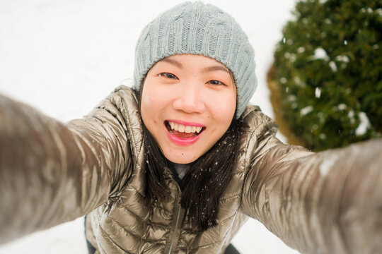 winter lifestyle portrait of young happy and beautiful Asian Japanese woman taking selfie picture with mobile phone enjoying snow at city park during Christmas holidays travel