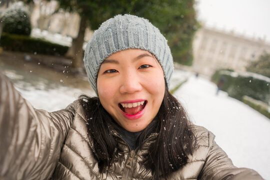 winter lifestyle portrait of young happy and beautiful Asian Korean woman taking selfie picture with mobile phone enjoying snow at city park during Christmas holidays travel