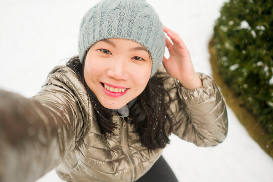winter lifestyle portrait of young happy and beautiful Asian Korean woman taking selfie picture with mobile phone enjoying snow at city park during Christmas holidays travel