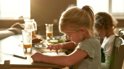 Portrait of focused pretty girl using tablet pc while having breakfast or lunch, sitting together with her brother at the table in the kitchen