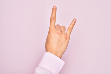 Hand of caucasian young man showing fingers over isolated pink background gesturing rock and roll symbol, showing obscene horns gesture