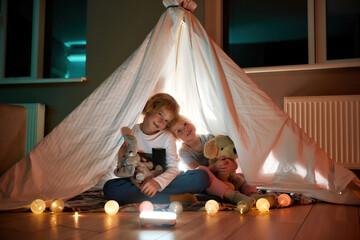 Happy children. Portrait of two little siblings smiling at camera, holding their toys while sitting on a blanket in a hut made with bedsheets