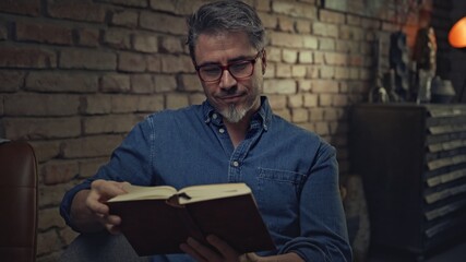Mature man in his 50s reading book at home sitting in dark living room.